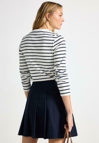 Stribet langærmet skjorte i creme og marineblå, parret med en marineblå plisseret nederdel. Outfittet har en tætsiddende talje og diskrete skulderknapper.