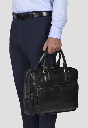 Porta documenti in pelle nera con doppie maniglie, una tasca frontale con zip e una finitura testurizzata, tenuto in mano da un uomo accanto a pantaloni blu navy.