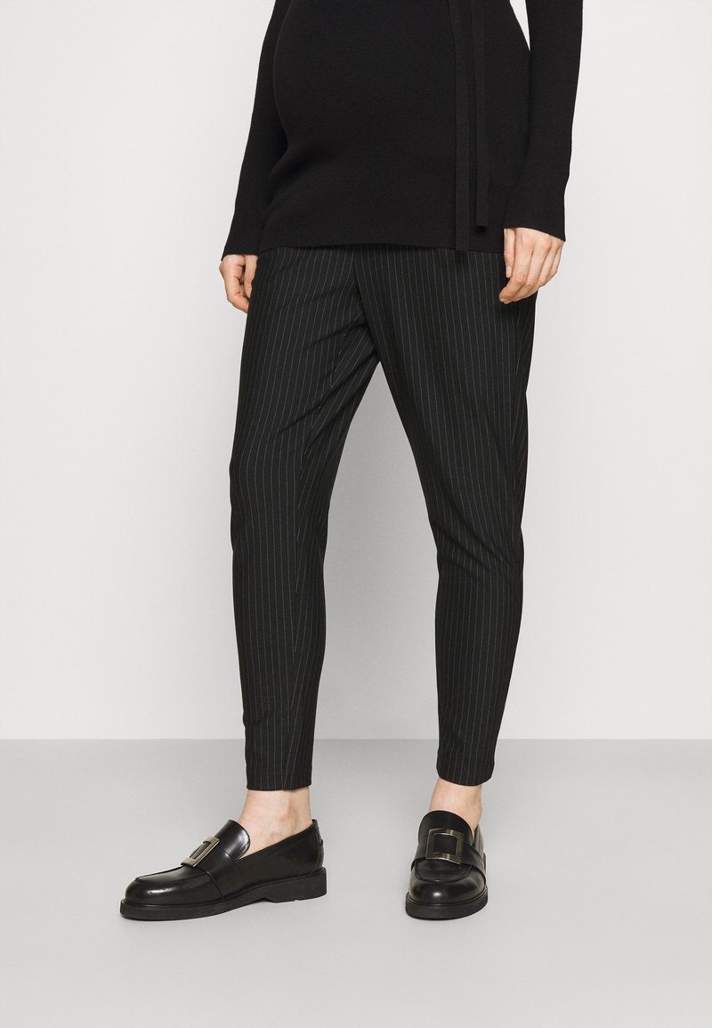 Personne portant un pantalon noir à fines rayures fuselé, des mocassins noirs avec des boucles argentées et un haut noir à manches longues, debout sur un sol gris.