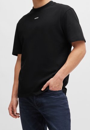 Homme portant un t-shirt Hugo noir uni à manches courtes avec un petit logo blanc centré sur la poitrine, associé à un jean bleu foncé.