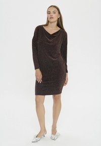 Moteris vilkinti kelių ilgio, ilgomis rankovėmis, tamsiai blizgančią suknelę su drapiruotu dekoltė ir sidabrinėmis žemais kulnais bateliais su didelėmis kaspinais.