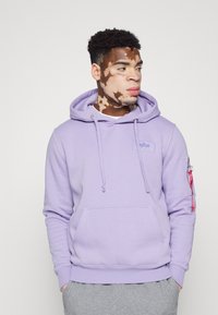 Lys lilla hættebluse med en frontlomme, justerbare snore og en sidelomme på ærmet med røde bånddetaljer. Blød tekstur.