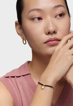 Donna con orecchini a cerchio d'oro e un braccialetto nero con ciondoli a forma di cuore in oro e argento, indossa un top rosa lavorato a maglia senza maniche.