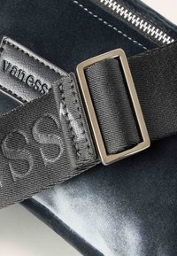 Bolso de terciopelo negro con cierre de cremallera plateada, una correa negra texturizada con texto en relieve y una hebilla metálica rectangular.