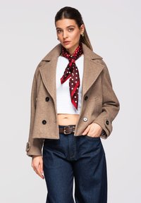 Žena nosící béžový vlněný krátký kabát, bílý krátký top, tmavé džíny, hnědý pásek a červený šátek s puntíky uvázaný kolem krku.