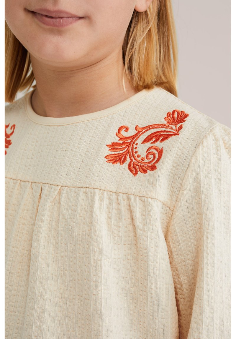 Blusa beige chiaro in tessuto testurizzato, con accenti floreali ricamati in rosso sulle spalle e un design arricciato sotto il collo.