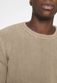 Pull en tricot beige avec un col rond, présentant un motif en côtes texturé et une coupe légèrement ample.
