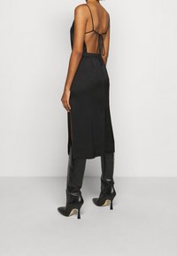 Robe en satin noir avec des fines bretelles, dos ouvert et fentes latérales. Portée avec des bottes stilettos noires hautes. Texture lisse, design minimaliste.