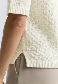 Maglione a maglia di crema texture con maniche corte, orlo a coste e fessure laterali, abbinato a pantaloni di colore chiaro.