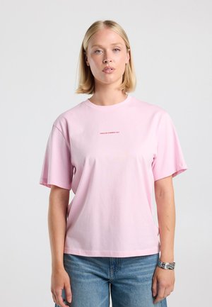 Blond vrouw met een lichtroze T-shirt met de tekst "ENDLESS SUMMER 1989" en een blauwe spijkerbroek, staand tegen een eenvoudige lichte achtergrond.