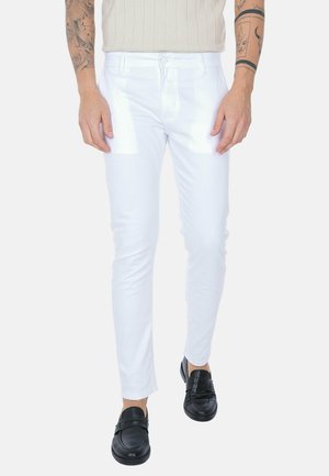 Uomo che indossa pantaloni bianchi aderenti, scarpe nere slip-on e una camicia beige a righe, con gli avambracci tatuati visibili.