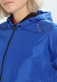 Niebieska wodoodporna kurtka z kapturem, zapinana na zamek, z czarnymi akcentami. Posiada logo "RLX" na piersi. Gładka, błyszcząca tekstura.