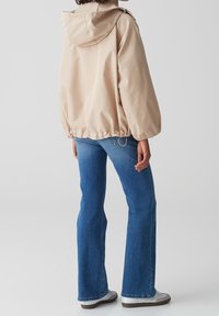 Persona che indossa una giacca a cappuccio beige, jeans a gamba dritta blu e sneaker bianche, in piedi contro uno sfondo grigio chiaro uniforme.