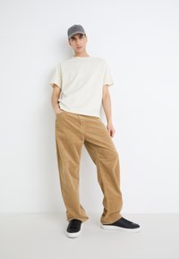 T-shirt en coton beige, coupe décontractée ; pantalon en velours côtelé marron, jambes larges ; baskets noires avec semelles blanches ; casquette grise. Design minimaliste et couleurs neutres.
