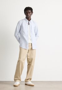 Mann står med hendene bak ryggen, iført lyseblåstripet skjorte med knapper, hvit undertrøye, beige bukser og hvite joggesko.