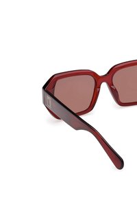 Ochelari de soare din acetat roșu cu un design hexagonal masiv, lentile maro, și un accent cu logo pe tâmplă. Textură netedă.