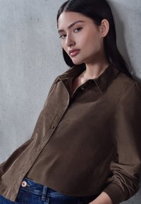 Brown, button-up shirt with a soft texture and a collar, paired with dark denim jeans. The shirt features a relaxed fit and long sleeves.
