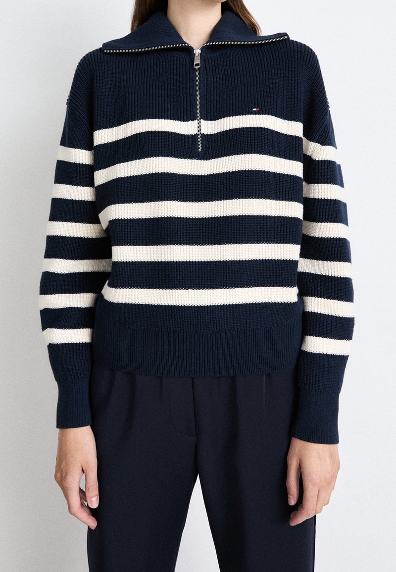 Mannequin portant un pull en maille bleu marine avec des rayures horizontales blanches et un col à demi-fermeture éclair, associé à un pantalon bleu marine.