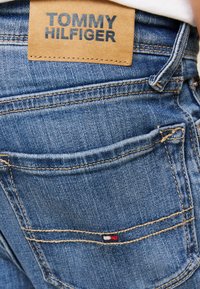 Plave traperice s istaknutom kožnom etiketom s natpisom "TOMMY HILFIGER" i malom logom u blizini stražnjeg džepa.