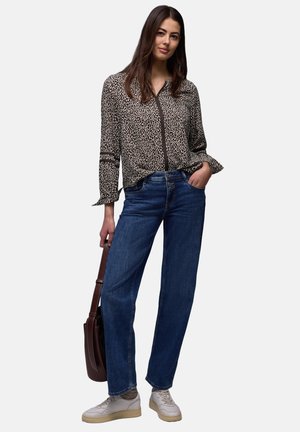 Femme portant un chemisier imprimé animal noir et blanc, un jean bleu, des baskets blanches, tenant un sac à main marron foncé avec une main dans la poche.