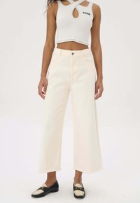 Witte wijde jeans met franjes aan de onderkant, hoge taille en zakken, gecombineerd met een nauwsluitend lichtgrijs mouwloos topje met een uitsnede.