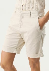 Beiga bomullsshorts med rak skärning, med sidofickor och uppvikta fållar, slät textur och sydda detaljer längs fållen.