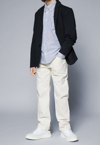 Blazer nero, camicia a righe blu, pantaloni crema, sneakers bianche. Il blazer ha rever; la camicia ha bottoni e colletto; le sneakers sono a basso profilo.