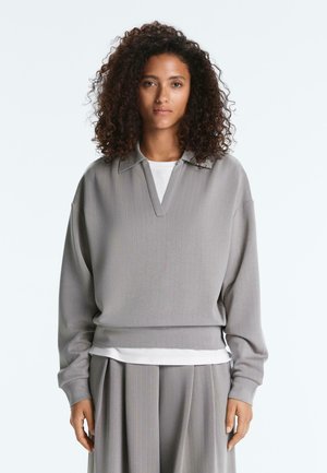 Frau mit lockigem Haar, die einen grauen, fein gestreiften V-Ausschnitt-Pullover über einem weißen Hemd und einem passenden grauen plissierten Rock trägt, steht vor einem schlichten Hintergrund.