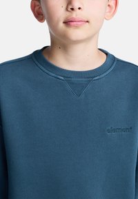 Modrá mikina so žebrovaným golierom a manžetami, s textúrovaným povrchom a vyšitým logom na hrudi. Materiál z bavlnenej zmesi.