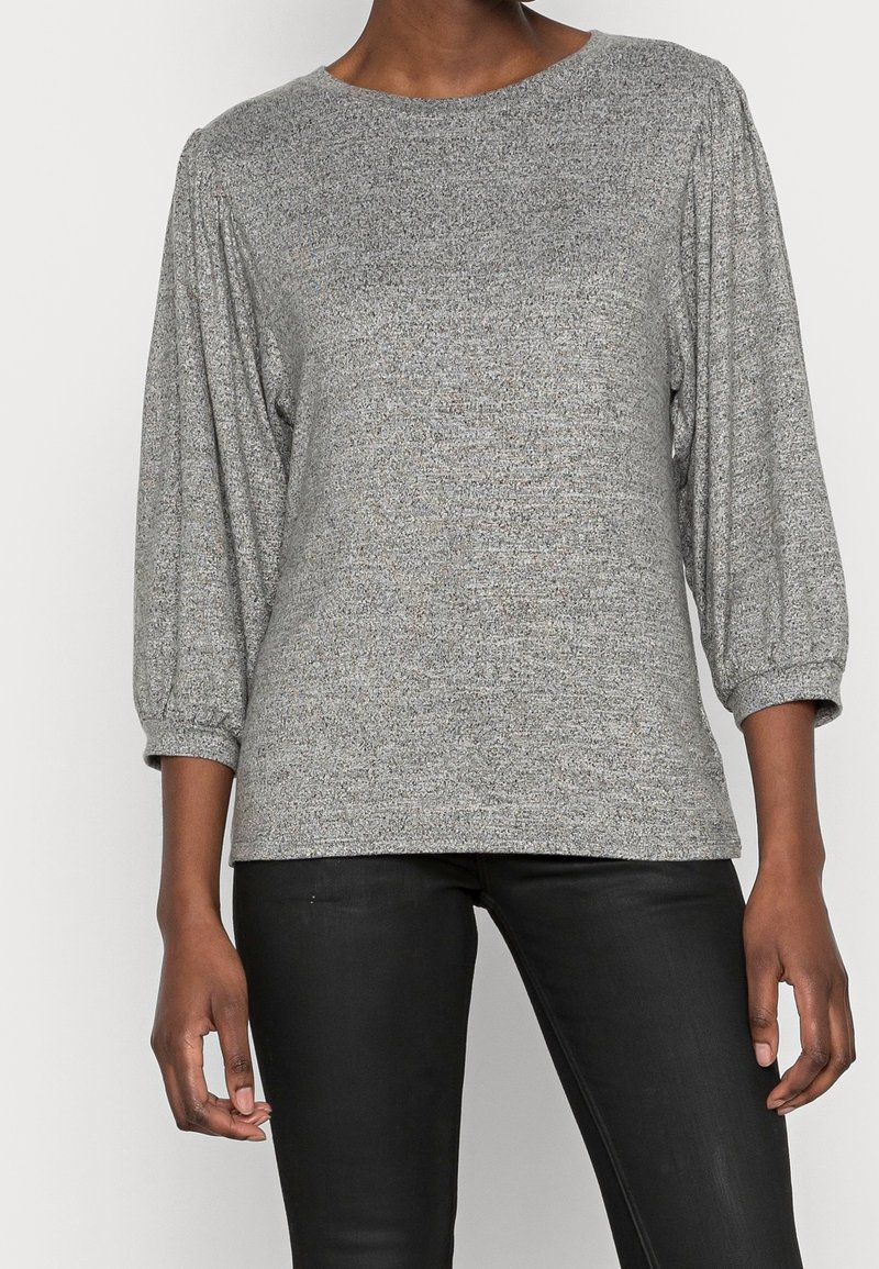 Pullover grigio texturizzato con maniche a tre quarti e scollatura rotonda. Il tessuto presenta un motivo sottile, abbinato a pantaloni scuri.