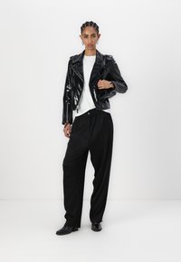 Veste de moto en cuir verni noir sur un t-shirt blanc, associée à un pantalon noir ample. Des chaussures noires étroites complètent le look.