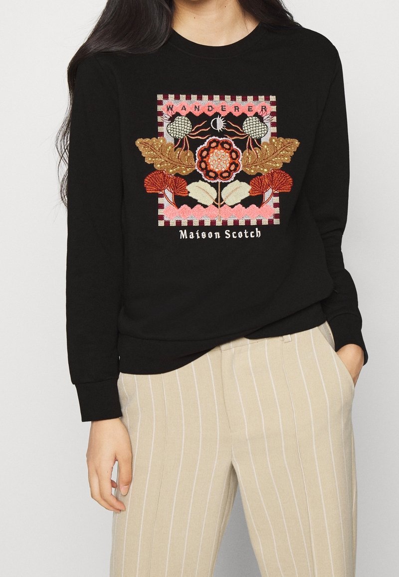 Sudadera negra con bordado floral multicolor, texto "WANDERER" y acentos de borde a rayas. Combínala con pantalones claros a rayas.