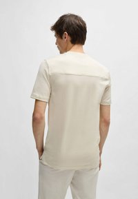 Camicetta corta beige chiaro in tessuto testurizzato con pannello posteriore liscio. Collo arrotondato, vestibilità ampia, abbinata a pantaloni chiari.