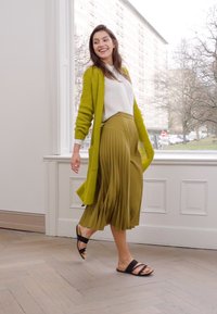 Cardigan vert citron tricoté au-dessus d'un top blanc, jupe olive plissée et sandales noires. Textures douces et formes fluides avec un arrière-plan en lumière naturelle.