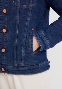 Person som bär en mörkblå denimjacka med bruna knappar, handen i framfickan, kombinerad med ljusbeige byxor mot en neutral bakgrund.