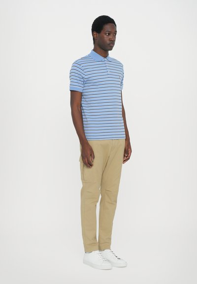 Μπλούζα πόλο με μπλε και λευκές ρίγες, κοντό μανίκι, σε συνδυασμό με καπαρντίνα χρώματος χακί και λευκά αθλητικά παπούτσια. Απαλή ύφανση και εφαρμοστή σχεδίαση.