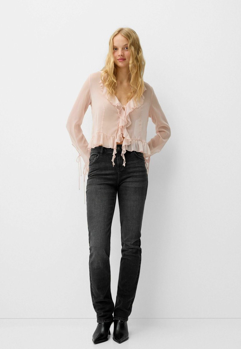 Donna che indossa una blusa rosa trasparente con balze, jeans neri attillati e stivali neri con punta a punta, in piedi contro un muro bianco uniforme.