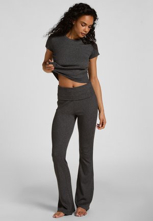 Vrouw met krullend haar, gekleed in een donkergrijs topje met korte mouwen en bijpassende wijde broek, waarbij ze haar shirt iets optilt om haar buik vrij te geven.