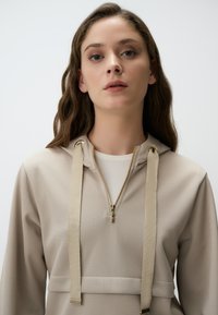 Felpa con zip beige in tessuto testurizzato, dotata di cappuccio, zip dorata e fasce piatte cucite per la regolazione. Design semplice, senza motivi.