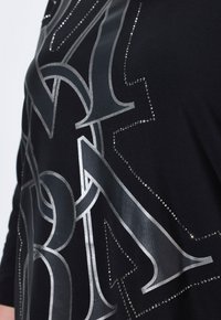 Schwarzer Stoff mit einem großen silbernen Grafikdesign, das metallische Akzente und Strasssteine aufweist, die die Formen und Muster umreißen.