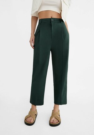 Femme portant un pantalon court taille haute vert foncé, des sandales beiges à brides croisées et un haut blanc court, debout devant un fond uni.