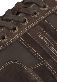 Braune Wildleder-Sneaker mit braunen Textilelementen, goldenen Ösen, kontrastierenden Nähten und einem Logo an der Seite.