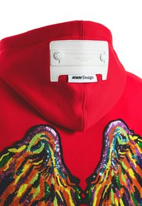 Sudadera roja con grandes diseños de alas de lentejuelas multicolores en la parte frontal, con una etiqueta blanca y botones en la parte trasera de la capucha.