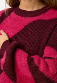 Adīts džemperis ar rozā un tumši sarkanvīna krāsu blokiem, teksturētu rakstu ar pūkainu diegu, ribotu kaklu un piedurknēm.