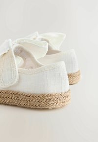 Espadrillas in tela bianca con suole in juta intrecciata e cinturini a fiocco in tessuto, viste da dietro su uno sfondo neutro.