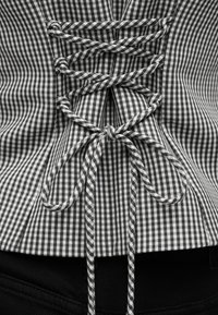 Μαύρο και άσπρο κορσέ τοπ με μοτίβο γκινγκχαμ, σφιχτά δεμένο με κορδόνια από το ίδιο ύφασμα, δεμένα σε φιόγκο στη μέση της πλάτης πάνω από μαύρο παντελόνι.