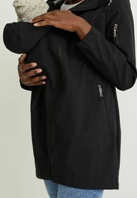 Musta softshell-takki, jossa on etuvetoketju ja sivutaskut, suunniteltu lapsen kantamiseen ja hupulla lisälämmikettä varten.