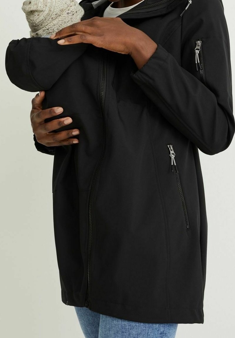 Musta softshell-takki, jossa on etuvetoketju ja sivutaskut, suunniteltu lapsen kantamiseen ja hupulla lisälämmikettä varten.