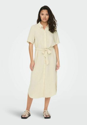 Light beige shirt dress with short sleeves, button-down front, tie waist, and knee-length hem. Paired with flat, strappy sandals.