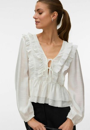 Žena s hnedým copom nosí bielu blúzku s dlhým rukávom s volánmi, výstrihom do V, viazankami vpredu a volánovým spodkom, v kombinácii s čiernymi nohavicami.